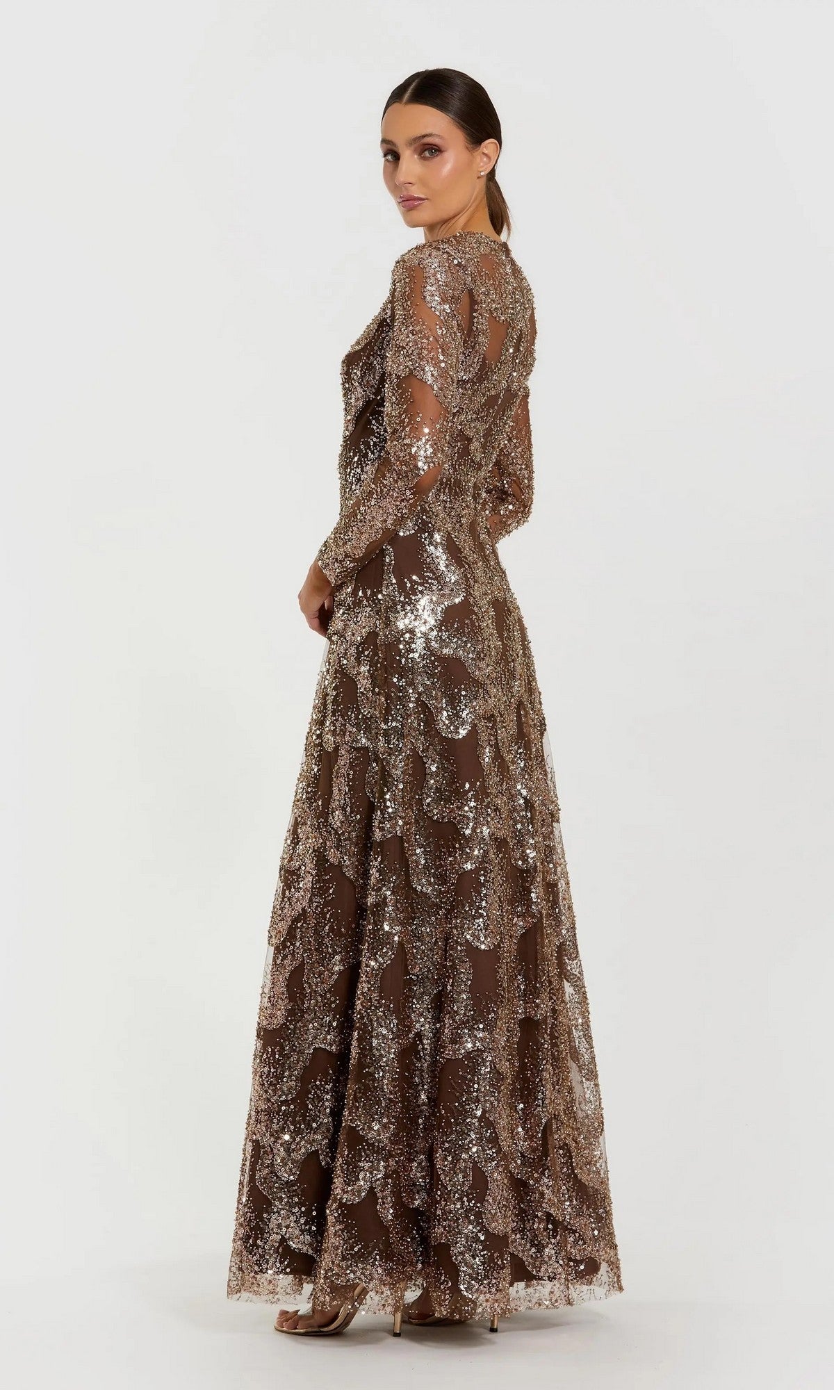 A woman wearing the Mac Duggal 69005 long formal dress—a brown, beaded sequin gown with sheer sleeves—stands against a plain white background, facing slightly to the side.